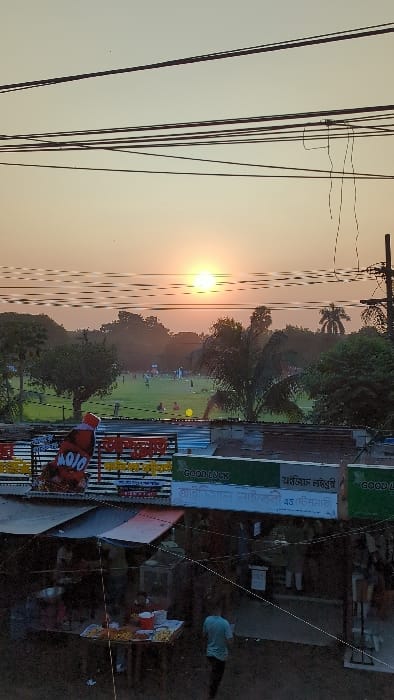 The image captures an urban landscape in Savar, Bangladesh, during either sunrise or sunset, indicated by a low, bright sun in a hazy, warm-toned sky. The foreground features a street-level commercial area comprising multiple stalls or small shops under corrugated metal roofs and fabric awnings. A food stall displays various prepared snacks on a table. One individual in a light blue shirt is observed walking away from the stall area on the right, with other indistinct figures present. Overhead signage includes a large "MOJO" beverage advertisement and green banners with the text "GOOD LUCK" in English, accompanied by Bengali script that partially translates to "Good Luck Library" and "Official Market and Stationery." Above this commercial layer, a expansive green field, appearing to be a park or sports ground, is visible. Multiple small groups of people are scattered across the field, some engaged in recreational activities resembling cricket or football, with a yellow spherical object (possibly a ball) on the grass. The field is bordered by numerous trees and foliage in the mid-ground and background. The upper section of the image is characterized by a dense network of dark electrical and utility wires, with a utility pole on the far right.