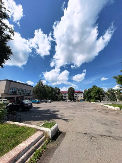 This is a medium-angle, wide shot of an outdoor parking lot on a bright, sunny day with scattered white clouds against a blue sky. The foreground features a cracked asphalt surface and a raised, concrete planter box with overgrown green grass and weeds. To the left, a dark-colored sedan is parked, and a weathered wooden bench is visible behind some green foliage.
In the midground, the parking lot extends towards a row of buildings and trees. A modern, light-colored building with large windows is situated on the left, possibly a cultural center or administrative building. Several cars are parked in front of it, including a black sedan, a bright blue hatchback, and other darker vehicles. Beyond these buildings, a residential apartment building with a red roof is visible, along with more mature trees lining the street.
To the far right, a metal playground structure and lampposts are present. The scene appears to be set in a town or city, with elements of both public buildings and residential areas. The overall impression is of a typical urban environment on a pleasant summer day, with no specific activity or event clearly depicted, suggesting it is an ordinary daytime scene. The presence of cars indicates normal daily operations for the buildings and surrounding area.
