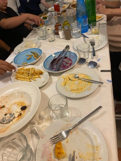 The scene depicts a dining table in a restaurant in Απολλωνία, Greece, following a meal. The table is covered with a white tablecloth, displaying numerous white and blue ceramic dishes. Most plates contain food remnants: a large oval dish holds traces of feta cheese, olives, and sauce; a round plate has mostly consumed yellow dip, likely hummus, with a spoon; another round plate shows pink/red and yellow residue with a single fried food item and a fork. A blue oval dish contains a few fried green vegetables, and a separate blue oval serving dish holds metal tongs in red liquid. A white round plate holds a visible portion of golden-brown French fries. Multiple other plates are mostly cleared. Beverages include two partially full glass bottles (one blue, one green, one labeled "ZAGORI") and a red Coca-Cola can, alongside several empty clear drinking glasses. Condiments such as oil, salt, and pepper are present in small glass bottles. A small container of bread is also on the table. A pair of light-colored sunglasses rests near the center. Used paper napkins are scattered on the surface. Parts of multiple diners are visible, including hands and forearms, indicating a recently concluded communal meal.