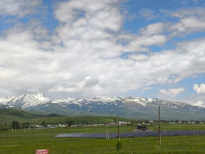 The image depicts a rural landscape in Apnagyugh, Armenia, featuring a vast green field in the foreground with a solar panel array stretching across it. In the middle ground, a village with various houses and structures is visible, nestled against rolling green hills. The background is dominated by a majestic mountain range with snow-capped peaks under a bright blue sky dotted with fluffy white clouds. The overall scene suggests a peaceful, agricultural setting with a touch of modern renewable energy infrastructure.