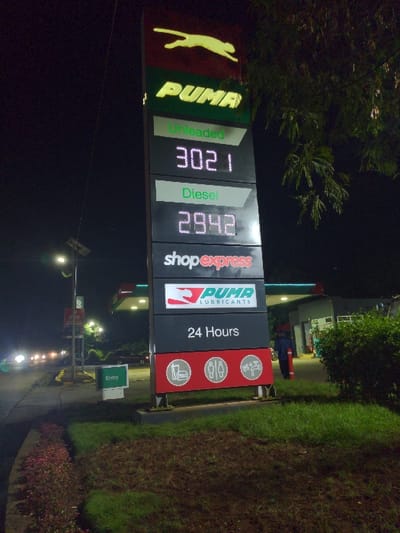 The image shows a Puma gas station sign at night. The sign displays the price of unleaded gas at 302.1 and the price of diesel at 294.2. The sign also indicates that the gas station is open 24 hours and has a shop express, and facilities for restrooms and car wash. A single individual can be seen walking on the asphalt of the gas station, away from the sign. The location is a gas station in Lindi Street, Tanzania, with coordinates -3.352 latitude and 37.338 longitude. The gas station is located in a well-lit area with lush green grass and trees. The ground appears to be asphalt, indicating a paved parking lot.
