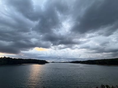 The image captures a body of water, likely a lake, framed by dense trees on either side. The sky is heavily overcast with dark, ominous clouds, giving the scene a somber mood. The water is calm with slight ripples, reflecting the subdued light from the sky. In the distance, land is visible, suggesting the far side of the lake or a small island. Based on the prompt, this scene is likely located in the city of Sugar Hill, United States.
