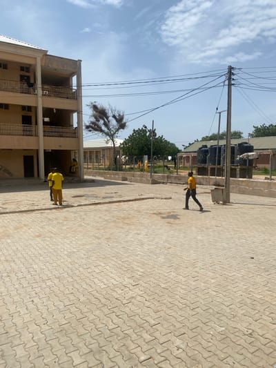This image depicts an outdoor scene in the city of Yan Dala, Nigeria. In the foreground, a paved area with interlocking concrete blocks leads towards a multi-story building on the left. The building appears to be made of concrete with cream-colored walls and has balconies with railings. Several individuals wearing yellow shirts and darker pants are visible in and around the building and the paved area. Two individuals are standing near the entrance of the building, engaged in conversation. Another person is seated on steps leading into the building. Further to the right, a single person in a yellow shirt walks across the paved area. In the background, there are other structures with corrugated metal roofs, fences, and trees. A utility pole with numerous wires is prominently placed in the middle ground, and several large black water tanks are visible behind a fence. The sky above is partly cloudy.
