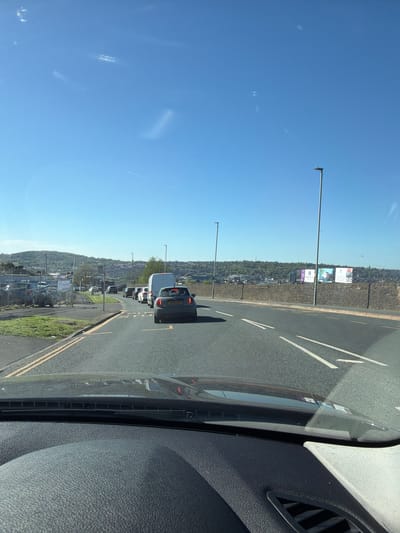 The image captures a scene from inside a vehicle, looking out through the front windshield onto a road in Blackburn, United Kingdom. The perspective is slightly angled, showing part of the vehicle's dark-textured dashboard and an air vent on the right.
Ahead, the road is visible with white dashed lane markings and double yellow lines along the curb. Several cars and a white van are in traffic, apparently stopped or moving slowly. The rear of a black car with the license plate 'AK08 RGO' is prominent in the mid-ground.
Beyond the road, a fence separates the road from a lower-lying area with parked cars. Further in the distance, a hillside rises, densely covered with trees and scattered buildings. Overhead, streetlights stand tall. On the hillside, several billboards are visible. One clearly displays "BLACKBURN College" with associated branding. Another billboard appears to advertise educational or vocational opportunities, possibly featuring text like "Future".
The sky is a clear, vibrant blue, indicating a bright, sunny daytime. A faint contrail from an aircraft is visible high in the sky. The overall impression is of a typical sunny day with urban traffic and a view extending to a green, built-up landscape.