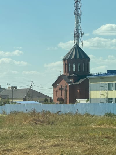 In the city of Shaumyanovsky, Russia, a cluster of buildings is visible under a bright, cloudy sky. The most prominent structure is a red brick church with a dark, multi-faceted dome topped with a cross. To the right of the church is a modern building with horizontal yellow and gray siding, featuring a visible external ladder and windows. A tall telecommunications tower stands behind and to the right of the church dome, with its metallic framework stark against the sky. In the middle ground, a blue-fenced area separates the buildings from a foreground of dry, tall grass and weeds. Other structures, including houses with dark roofs and power lines, are visible in the background.
