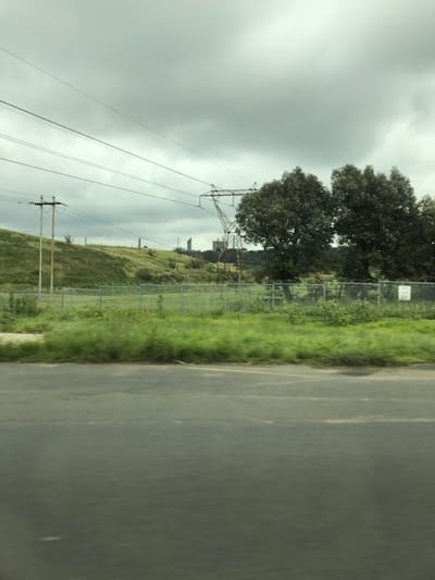The image depicts an outdoor scene in Kempton Park, South Africa, under a uniformly overcast sky. In the immediate foreground, a dark grey asphalt road surface, possibly damp, spans the width of the frame, with faint indications of a yellow line along its distant edge. A strip of green grass and dense vegetation, varying in height, runs parallel to the road. Behind this vegetation, a chain-link fence with metal posts stretches horizontally across the midground, partially obscured by foliage.
Beyond the fence, a expansive green field or open grassy area is visible, sloping gently upwards towards the left. On the right side of the midground, two large, mature trees with dense, dark green foliage stand behind the fence. Further back, on the left, a weathered wooden utility pole with cross arms and multiple overhead lines rises from a grassy incline.
A prominent lattice steel electricity pylon with numerous transmission lines spans across the mid-background, connecting to the upper left. In the distant background, atop an undulating green hill, several smaller, indistinct utility poles or structures are scattered. On the distant horizon, industrial structures are visible, including a tall, slender, light-colored cylindrical tower and a larger, multi-level industrial building or plant complex. No human figures, animals, or active subjects are present in the scene.