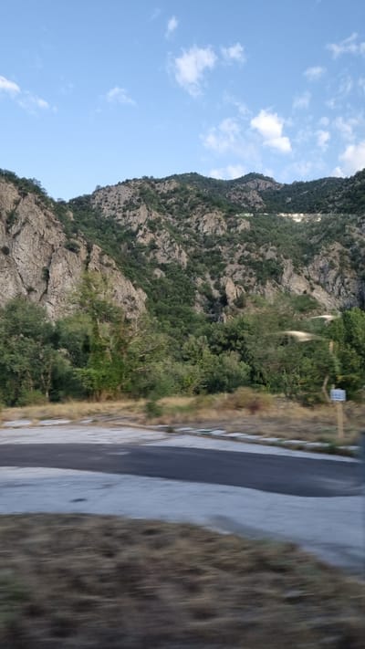 The image depicts a mountainous landscape with a road in the foreground. The mountains are rugged and covered with green vegetation, primarily trees and shrubs. The sky is blue with scattered white clouds. The road appears to be paved and has a light-colored border. There is dry grass and some smaller plants along the side of the road. A thin, dark pole with a white sign is visible on the right side of the image. The image appears to have been taken from a moving vehicle, as there is motion blur in the foreground, particularly in the dry grass. The overall impression is of a natural, somewhat wild environment, potentially on the outskirts of a town or city. Based on the context provided, this scene is located in Kresna, Bulgaria.