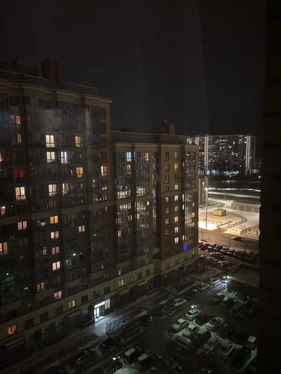 A night view from a high vantage point showcases a tall, multi-story apartment building with numerous illuminated windows. A brightly lit parking lot filled with closely packed cars stretches to the building's right. In the distance, another high-rise structure glimmers with lights. The sky is dark, hinting at the late hour. The image is shot at a slightly tilted angle, capturing the urban environment's density.