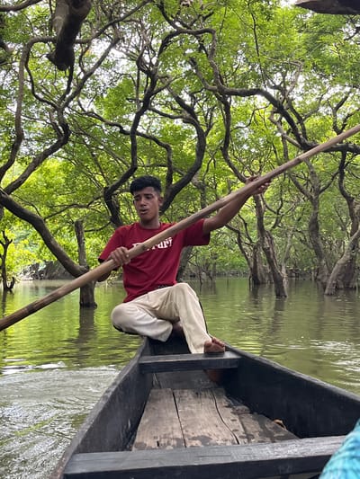 A young man is rowing a boat through a waterlogged forest, likely the Ratargul Swamp Forest near Sylhet, Bangladesh. He is wearing a red t-shirt and light-colored pants, and is seated on the floor of the wooden boat. The water is green and reflects the dense foliage of the trees that rise from it. The branches of the trees intertwine overhead, creating a canopy that filters the sunlight. The man is holding a long wooden oar, which he is using to propel the boat. The scene is serene and idyllic, with the only sounds being the gentle lapping of water and the rustling of leaves.