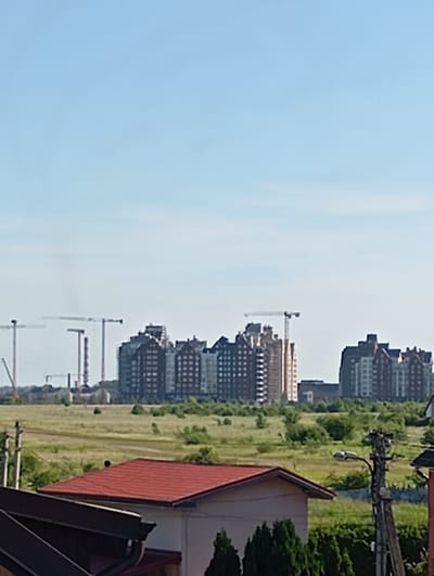 The image depicts a suburban or semi-rural landscape with residential buildings in the foreground and mid-ground, and a cluster of larger apartment buildings and construction cranes in the background. The sky is clear and blue, indicating daytime.
In the immediate foreground, the roofs of two houses are visible. The house on the left has a dark, angled roof, possibly made of metal or asphalt shingles. The house on the right has a bright red, sloped roof. A portion of a light-colored wall is visible on the right house. Adjacent to this house, a wooden utility pole with a street lamp is prominent.
The mid-ground is characterized by a field of tall, dry grass with scattered bushes and trees. A dirt road or path appears to run through the field.
In the background, a line of multi-story apartment buildings is visible. These buildings have a modern architectural style, with numerous windows. To the left of the apartment buildings, several construction cranes are erected, suggesting ongoing development in the area. One crane is particularly tall and appears to be actively lifting materials.
The overall impression is of a developing residential area, possibly on the outskirts of a city, with a mix of older, single-family homes and new, multi-unit dwellings under construction. The environment appears to be a green, open space surrounding the developed areas. The specific location of Maloye Isakovo, Russia, is implied, suggesting this scene is from that region.