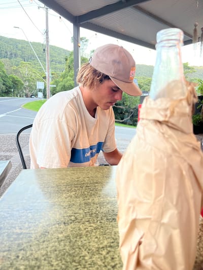 The scene is an outdoor dining area in Stanwell Park, Australia. A young adult male is seated at a speckled green and black table, positioned left of center. He has light skin, visible shaggy light brown hair, and a light mustache. He wears a beige baseball cap featuring a square patch with a colorful graphic, and a light beige t-shirt with a horizontal blue stripe across the chest, containing the word "Billabong". His gaze is directed downward and to his right, towards the table surface. In the immediate foreground, to the right, a clear glass bottle with a metallic cap is largely obscured by crinkled light brown paper or plastic wrapping. The background reveals a paved road with white markings, a wooden utility pole with wires and a street light, and dense green, forested hills. Overhead, a corrugated awning with decorative string lights is visible. The lighting is soft, consistent with late afternoon or early evening.