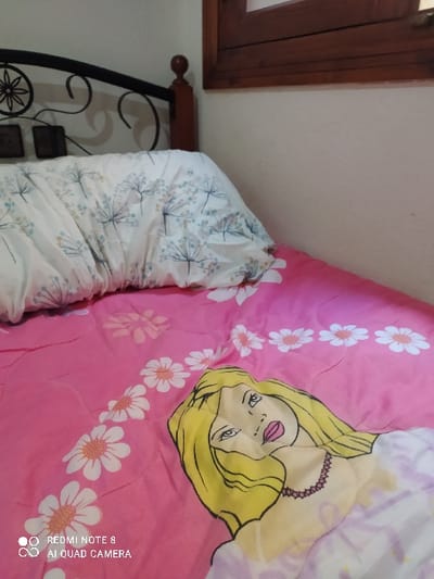 The image depicts an indoor bedroom scene located in Tétouan, Morocco. A bed is centrally featured, presenting a dark brown wooden and black wrought iron headboard with decorative scrollwork. A white pillow with a light blue and green dandelion-like floral pattern rests against the headboard. The bed is covered with a pink comforter. This comforter displays a repeating pattern of white daisies with orange centers, arranged in diagonal stripes, and a large graphic illustration of a blonde-haired female character wearing a pink necklace. The background consists of a light-colored wall, with a portion of a dark brown wooden window frame visible in the upper right corner. No individuals, actions, or interactions are present in the scene.