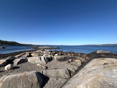 This wide shot captures a serene coastal landscape at Nærsnes, Norway, under a vast, clear blue sky. The foreground is dominated by large, grey, weathered rocks of varying sizes and shapes, interspersed with smaller gravel and pebbles. Shadows on the rocks suggest bright daylight, likely late morning or early afternoon.
The rocky shoreline extends into the midground, meeting the calm, dark blue water of a fjord or bay. The water appears still, with a few faint ripples, and reflects the clear sky subtly. In the distance, on the left side of the frame, a densely forested hillside rises from the water's edge, with some structures visible among the trees. Further along the distant shore, a distinct cluster of cylindrical industrial buildings, possibly cooling towers or silos, is visible.
To the right, distant landforms, likely islands or the far side of the fjord, stretch towards the horizon. A few tiny white specks on the water indicate distant boats, possibly sailboats. The sky is an unbroken expanse of brilliant blue, transitioning from a slightly darker shade at the top to a lighter hue near the horizon, indicating cloudless and fair weather. The scene is devoid of people or direct activity, conveying a sense of peace and natural grandeur. No visible text is present in the image.