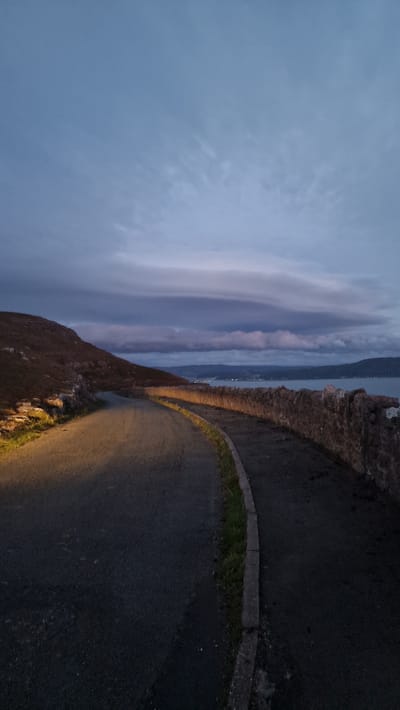 The image depicts a coastal road at dusk or dawn. The road curves to the left, with a low stone wall bordering the right side of the road. Beyond the wall, a body of water stretches to the horizon, with hills and lights visible in the distance on the far side. A steep, grassy hill rises on the left side of the road, with rocky outcrops visible. The sky is overcast with dramatic clouds. The lighting suggests a time of low light, possibly dawn or dusk, with artificial lights illuminating a portion of the road on the left. There are no people or vehicles visible in the scene. The location is not specified in the image itself.