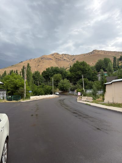 The image depicts a road leading towards a mountainous backdrop in Khumsan, Uzbekistan. The road is paved and appears wet, with reflections visible on its surface. On the left side of the road, there is a white car partially visible, along with a green metal fence and a concrete structure. Further down the road, there are utility poles with electrical wires. Trees and vegetation line the sides of the road, and in the distance, a hillside is visible, sparsely covered with dry grass and scattered bushes. The sky is overcast with heavy grey clouds. A sign with "HUMSON VILLA" written on it is visible near a utility pole. Buildings with simple architecture are present among the trees. The overall scene suggests a rural or semi-rural area on a cloudy day.