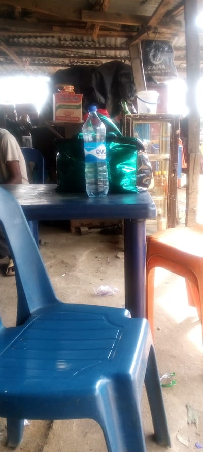 The image shows a scene in Elelenwa, Nigeria, taken under a corrugated metal roof. A blue plastic chair sits in the foreground, positioned at a table with a blue top and blue legs. A blue plastic water bottle, a green bag, and a box of noodles are on the table. An orange plastic chair is partially visible in the background. A person wearing a white shirt is sitting in a blue chair, but they are only partially visible. The image was taken in an outdoor space, likely a food stand. The location is determined by the coordinates provided.