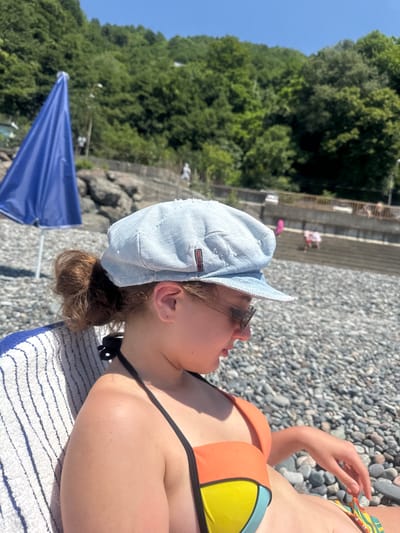 The media file captures a close-up of a young person lying on a towel on a rocky beach. The person is wearing a light blue denim cap and sunglasses, with their head tilted upwards and to the right, looking towards the sky. Their hair is pulled back into a bun. They are also wearing a colorful bikini with orange, yellow, and blue sections. A portion of a blue umbrella is visible in the lower left corner of the frame.
The background suggests a sunny day at the beach, with lush green trees on a hillside visible behind a retaining wall. The ground is covered in small, smooth pebbles. The lighting indicates it is likely daytime, possibly midday given the bright sunshine. The overall mood appears relaxed and summery.
A small tag on the denim cap is visible, although the text is difficult to read clearly. It appears to be a brand logo, possibly with some letters like "ON" and "BE".