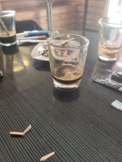 A black table top with diagonal striations is the surface of a cafe table in Tunis, Tunisia, located at 36.838 latitude and 10.128 longitude. On the table are three glass cups. Two of the cups are empty, one with a slight residue at the bottom. The third cup is partially filled with a dark liquid, likely coffee, and is situated near the center of the table. To the right of the cup, a small black rectangular box with gold lettering lies flat on the table. Next to the box is a small white object, perhaps a piece of plastic. A brown ashtray rests on the table, left of the cup of coffee. The ashtray appears to have some cigarette butts, or similar, debris inside. Two small wooden objects resemble pieces of a broken match or stick lying on the table, in the lower left of the photo. The left-hand side of the table's edge is partially obscured by a black object, likely a chair or a part of a table. The background of the photo is out of focus. It appears to be a wooden surface. There is no sign of people in the photo. The scene suggests a quiet cafe, perhaps after a recent patron left.