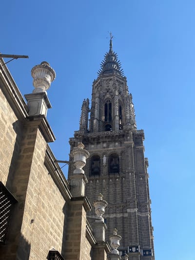 The image displays the bell tower of the Primate Cathedral of Saint Mary of Toledo, located in Toledo, Spain, under a clear blue sky. The tall, ornate Gothic tower is constructed from grey stone, featuring multiple levels with arched openings, some containing bells, intricate tracery, statuary (including two white figures in arches), and decorative pinnacles, culminating in a spired lantern topped with a finial. In the foreground, positioned to the left, is a section of a light tan, rectangular stone block wall, likely part of an adjacent structure or the cathedral complex. This wall features a prominent parapet adorned with multiple white, classical-style decorative urns. Sunlight illuminates the scene, casting defined shadows on the foreground wall. No individuals are visible.