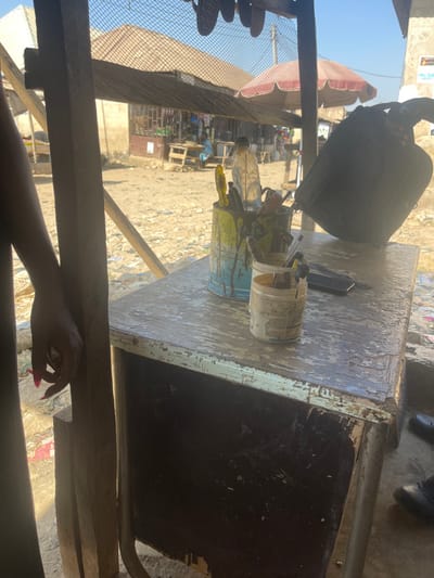 This image depicts an outdoor market or street stall scene in Dzhidu, Nigeria, under clear daylight conditions.
In the foreground, a person's left forearm and hand are visible on the far left, resting against a wooden support beam. The hand features long, pointed, bright pink fingernails and appears to be loosely holding a small, indistinct object. Adjacent to this, a worn wooden table occupies the central-right foreground. Its surface is light-colored, showing peeling paint and various dark and light stains, including drips of paint. On the table, two repurposed containers hold tools: a larger blue and yellow paint-splattered metal can contains several screwdrivers, a clear plastic water bottle, and other elongated implements. A smaller, white jar, stained with orange/brown residue, sits to its right, containing additional small tools, possibly brushes or picks. A dark wallet or small rectangular object and a pen are also present on the table surface. A dark bag or backpack hangs from a support structure to the upper right of the table. Partially visible at the bottom right are dark shoes, indicating the presence of another individual near the table. Overhead, a mesh net structure is visible, supported by wooden beams.
The background reveals a dusty, unpaved street or open area with scattered debris. Several single-story buildings constructed from light-colored materials are visible, some with corrugated metal roofs. Market stalls with wooden tables are discernible, and a red and white striped umbrella provides shade over one area. Two distant individuals are observed: one seated at a stall in a light blue garment, and another standing further down the street. The sky is clear and blue, indicating strong sunlight casting distinct shadows.