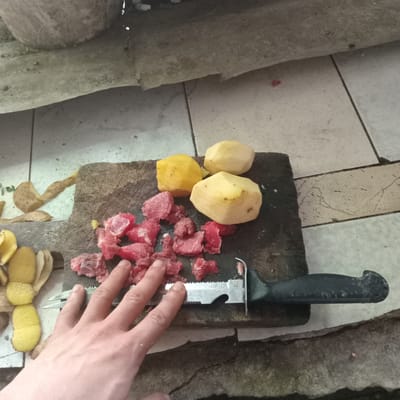 The image shows a close-up overhead view of a person's hand reaching over a cutting board. On the cutting board are several peeled potatoes and a pile of chopped raw red meat. A serrated knife with a black handle is also on the board, partially under the hand. The cutting board is rustic and appears to be made of wood. The background shows a tiled floor and what looks like a concrete structure. The lighting suggests it could be daytime, but there are no strong indicators of the weather. The scene appears to be in a kitchen or an outdoor food preparation area. The focus is on the food preparation, suggesting a meal is being made. There is no visible text in the image. The location context provided is Duaca, Venezuela.