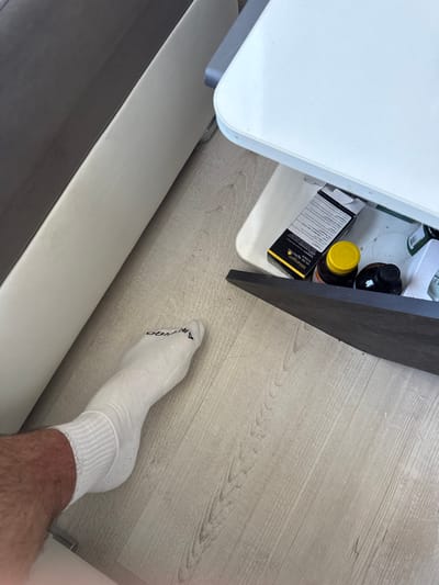 A person's left leg, wearing a white sock with a black logo, is visible in the foreground. The leg is positioned on a light-colored wooden floor. Behind the leg, a white surface, possibly a table or desk, extends into the background. Partially opened, a drawer or compartment beneath this surface contains several items: a black and yellow box, a bottle with a yellow cap, and a dark bottle. The environment appears to be indoors, with the lighting suggesting daytime. There is no discernible information about the location in the city of Dibich, Bulgaria.
