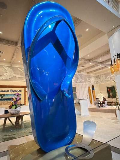 This is a wide shot of a large, shiny blue flip-flop sculpture in a brightly lit, modern lobby. The sculpture is the primary focus, dominating the foreground and mid-ground. In the background, there is a seating area where two people are seated at a table. There are also surfboards displayed on the wall, a painting of a parrot, and decorative wall art depicting birds and palm fronds. The ceiling has a coffered design with recessed lighting. The scene appears to be located in a hotel or resort lobby, possibly in Hollywood, Florida, given the tropical-themed decor.