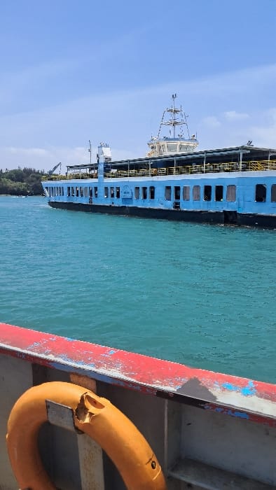 The image displays a daytime maritime scene in the Ganjoni/Shimanzi ward, Kenya, featuring a large, light-blue multi-deck ferry. The ferry occupies the midground, characterized by a dark hull, numerous rectangular windows along its side, and an upper deck with a canopy. A prominent white superstructure, housing a bridge with communication masts and antennae, rises from the vessel. The body of water is turquoise, indicating clear conditions. In the background to the left, a green, tree-lined landmass is visible, featuring a large industrial crane. The foreground provides a partial view of another vessel, from which the photograph was taken, showing a weathered red and blue painted railing with visible paint chipping. An orange lifebuoy, distinctly damaged with a significant crack on its upper surface, is mounted to this railing on the lower left. No individuals are present in the frame. The large ferry appears to be afloat, positioned, or in transit within the channel.