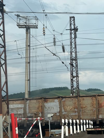 This image shows a train at a railway crossing on a cloudy day in Zykova, Russia. The train cars are weathered and rusty, with Cyrillic text and numbers visible on their sides. A railway crossing gate with red and white stripes is positioned in the foreground. Overhead, a complex network of electrical wires and insulators, supported by concrete and metal poles, spans across the frame. In the background, rolling hills covered in lush green trees are visible under a grey, overcast sky. The scene depicts typical infrastructure and landscape of a rural railway in Russia.