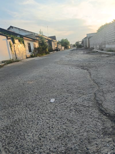 The image depicts a street scene in Yangi Chinaz, Uzbekistan, during what appears to be late afternoon or early evening, given the soft, warm lighting. The sky is partly cloudy with a hazy sunset glow, suggesting clear weather. The street itself is paved with asphalt that shows signs of wear and tear, with visible cracks and an uneven texture.
Along the left side of the street, there are several low-rise buildings. One building prominently features large metal garage doors, some of which are illuminated by the golden sunlight, casting long shadows. Green vines climb the wall of this building. Further down the street, other structures with windows and some vegetation are visible. On the right side of the street, a long, high wall constructed from grey concrete blocks runs along the entire length of the visible area. Beyond this wall, more buildings and trees can be seen in the distance.
In the foreground of the street, a small piece of white paper lies on the asphalt. The scene is quiet and appears to be a residential or semi-industrial area. There are no people visible in the image. A small, dark-colored vehicle is parked on the left side of the street further down. The overall impression is of a slightly weathered but functional urban environment at the end of the day. There is no visible text in the image.