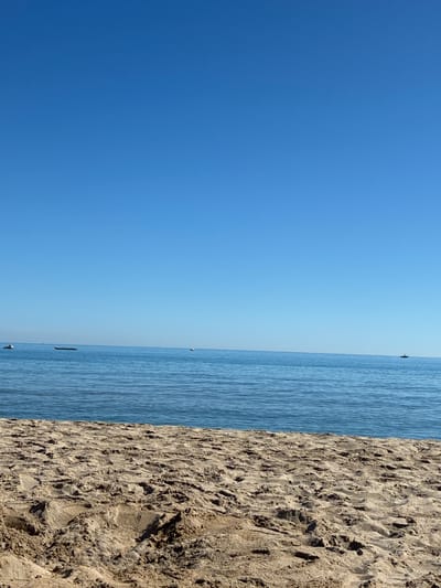 The image shows a clear, bright, and sunny day at a sandy beach. The foreground is dominated by the coarse texture of dry sand, with visible footprints and undulations indicating recent activity. Beyond the sand, a calm, blue sea stretches out to the horizon, meeting a vast, cloudless sky of a similar cerulean hue. Several small objects are visible on the water's surface: a jet ski on the far left, a long, dark, flat object that could be a buoy or a small raft in the middle distance, and a dark, distant boat to the right. The overall impression is one of a serene, idyllic beach scene.
This scene is located on the coast of Rethymno, Greece.