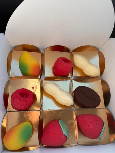 A white cardboard box, open and viewed from an overhead perspective, contains nine individual dessert confections arranged in a 3x3 grid within golden, square-shaped paper liners. This scene is situated in the city of Cesena, Italy. The top row features, from left to right, a mango-shaped confection transitioning from green to yellow, a red raspberry-shaped confection, and an off-white peanut-in-shell-shaped confection. The middle row displays another red raspberry-shaped confection (with a small red stain on its liner), another off-white peanut-in-shell-shaped confection, and a dark brown coffee bean-shaped confection. The bottom row includes a second mango-shaped confection, a red strawberry-shaped confection with a green leaf, and another red strawberry-shaped confection with a green leaf positioned along its side. The inside of the box is white, and the upper flap reveals faint, light-colored text and a small leaf-like logo. The external background is dark and out of focus.