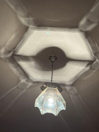 An antique chandelier hangs from a ceiling, casting intricate shadows. The ceiling has a hexagonal recess with a light fixture at its center. The chandelier itself is made of glass panels held together by a metal frame, with a single light bulb illuminating the space. The shadows projected onto the ceiling are a prominent feature of the image, creating a dramatic and somewhat abstract pattern. The overall scene is dimly lit, with the chandelier being the primary light source. The location is specified as Krumovgrad, Bulgaria.