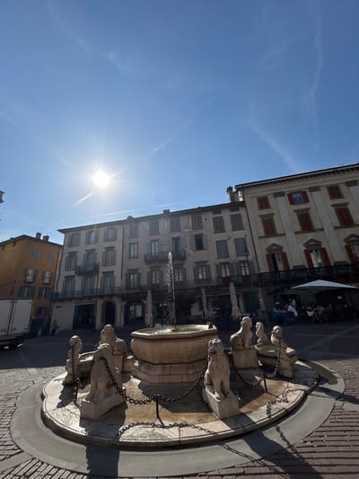 In Piazza Matteotti, Bergamo, Italy, a sunlit scene unfolds with a circular fountain at its center, adorned with lion sculptures and chains. Buildings with red-shuttered windows flank the square, reflecting the bright sky. People are seated at tables under umbrellas, enjoying the day. A delivery truck is parked to the left of the frame. The sun's glare is strong, casting sharp shadows across the cobblestone pavement.