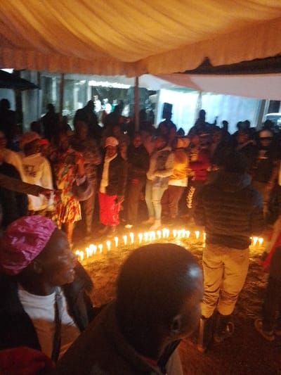 The image depicts an outdoor nighttime gathering under a light-colored fabric canopy in Marenyo-Shianda ward, Kenya. Numerous individuals are present, standing and seated, with a prominent circle of approximately 50-60 lit white pillar candles arranged on the dark, possibly earthen, ground in the midground. The primary light source appears to be the candles and ambient lighting from overhead within the canopy, casting warm tones.
In the immediate foreground, the left side shows the profile of an individual with dark skin, wearing a pink, possibly satin, head covering and a white shirt. To the right of this person, the bald head and upper back of another individual are visible, facing away from the viewer.
A dense crowd of people occupies the midground, surrounding the candle circle. Their attire is casual and varied, including long skirts, jeans, hoodies, and patterned tops. Many faces are obscured by low light, distance, or partial views. In the background, indistinct figures are visible under further tent structures, one of which appears to have a blue-tinged interior light. A dark, rectangular object, possibly a speaker on a stand, is visible on the right side of the background. No specific interactions are clearly discernible within the crowd; individuals appear to be observing or participating in the event.