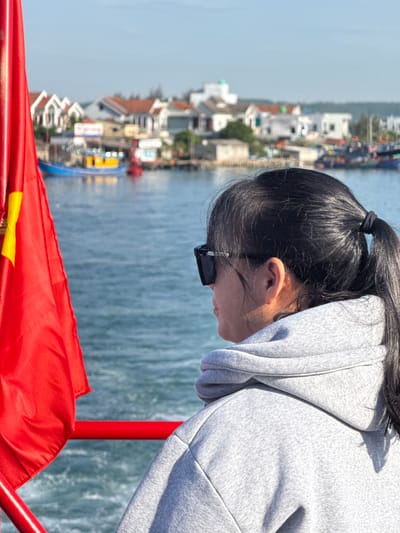 The image depicts a woman from the chest up, viewed from behind and slightly to the side, facing a body of water. She has dark hair pulled back in a ponytail and wears sunglasses and a grey hooded sweatshirt. A red Vietnamese flag with a yellow star is prominently displayed on the left side of the frame, partially obscuring the background. In the distance, across the water, a coastal town is visible, characterized by rows of houses and several boats docked or moored. The scene suggests a location along the coast of An Hải, Vietnam, possibly a harbor or a waterfront area. The water is choppy, indicating movement, and the sky is clear and bright. The red railing in the foreground suggests the woman is on a boat or a pier.