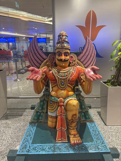 The central subject of the image is a brightly colored, intricately detailed statue, likely depicting Garuda, a divine bird-like creature from Hindu mythology. The statue features a human-like torso with a mustache-adorned face and large wings, rendered in vibrant shades of yellow, orange, red, green, and blue. It is adorned with elaborate jewelry, headwear, and patterned garments. The figure stands on an ornate, rectangular base painted in green and blue.

The scene is set indoors, likely within a modern airport terminal or a similar large public building. In the immediate background to the left, a large, stylized orange and blue logo resembling a bird or a leaf is displayed on a white wall. A potted plant is visible at the top edge of the frame. Further into the background, the polished floor reflects the surroundings, and what appear to be check-in counters or information desks with blue screens are discernible, hinting at a bustling public space.

No specific activity or event is taking place; the image captures a static display. The lighting is artificial and bright, indicating an indoor environment. A notable detail is a symbolic emblem, possibly a chakra, visible on the statue's raised right palm. The statue's expression is stern and majestic. Given the location context of St. Thomas Mount Cantonment, India, the setting is consistent with Chennai International Airport (MAA). No other clear, readable text is present in the image.
