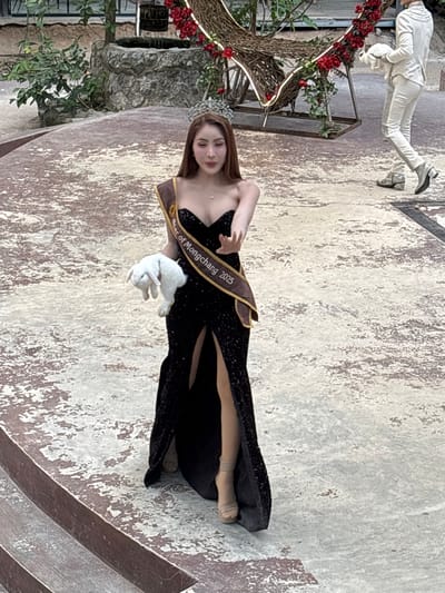 A woman wearing a black sequined gown, a crown, and a sash that reads "Star of Mongchang 2025" holds a white rabbit in her left hand. She is standing on a concrete-like surface with a distressed, mottled pattern. To her right, another person in a white suit walks away from the camera, also holding a white rabbit. In the background, there is a decorative structure adorned with red flowers and lights, a stone well, and lush green foliage. The scene appears to be outdoors, possibly in a garden or courtyard setting within the city of Nong Prue, Thailand.