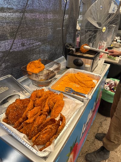 A food stall in Mazkeret Batya, Israel, located at coordinates 31.833, 34.82, features deep-fried chicken strips coated in sesame seeds, displayed in a metal tray lined with white paper. The chicken is in a state of readiness for sale. Additionally, there are two large metal trays containing more fried chicken, one of which is being prepared in a deep fryer with the assistance of tongs. A large metal fan is positioned behind the food preparation area, suggesting an attempt to control temperatures and ventilation. A person wearing brown cargo pants is visible, standing behind the food stall and reaching into the deep fryer, while their legs are also seen below. There is a black trash can containing various items to the right of the stall. The stall itself is painted with a bright blue surface featuring a cartoon image of a red strawberry. The background consists of a black metal mesh wall.