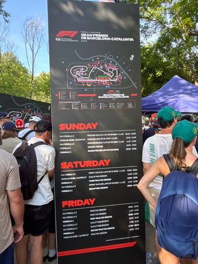The image shows a close-up view of a race schedule sign in Montmeló, Spain, likely at the Circuit de Barcelona-Catalunya. The sign, predominantly black with red and white text, displays the schedule for Formula 1 and other racing series for Sunday, Saturday, and Friday. The schedule includes details for practice sessions, qualifying sessions, and races, along with their respective times.
In the foreground, several people are visible from behind, wearing casual clothing and some sporting racing-themed caps and backpacks. A blue backpack is prominently featured on the right side of the image. The background is somewhat blurred, showing trees and a glimpse of a blue tent or canopy. The overall scene suggests an outdoor event with attendees viewing the information board.