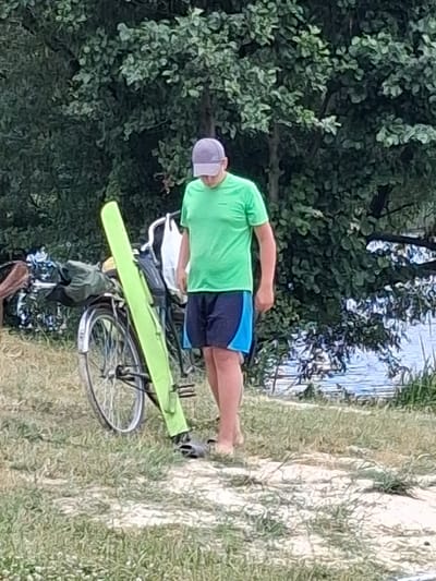 A person wearing a green t-shirt and blue shorts is standing next to a bicycle. The person is wearing a light-colored cap. The bicycle has a bright green bag or case attached to its frame. The scene appears to be outdoors, with lush green trees and vegetation in the background, and a body of water visible behind the trees. The ground is covered in grass and what looks like a dirt path. The lighting suggests it is daytime, possibly late afternoon, given the angle of the shadows. The overall impression is of a casual, outdoor recreational setting. The image is oriented vertically, with the person and bicycle appearing to be on the left side of the frame, and the natural landscape on the right. The location is identified as Reutyntsi, Ukraine. No text is visible in the image.