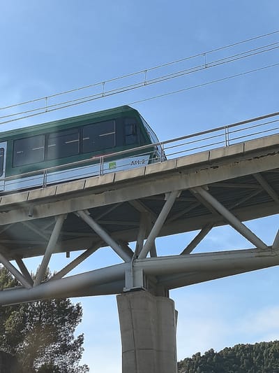 The image captures an elevated green and silver train, resembling a monorail or a rack railway car, positioned on a substantial concrete and steel bridge structure against a bright, clear blue sky. The train features multiple dark, rectangular windows along its side and has the alphanumeric identifier "A11.2" along with a small green logo visible near its front. Overhead, several parallel power lines run above the track.
The train is supported by an elevated concrete track bed, which includes a silver metal railing. Beneath this, a robust grey steel truss structure forms the bridge's main support, featuring numerous interconnected beams and diagonal braces. This steel framework rests on large, sturdy concrete pillars.
In the background, under the bridge and extending towards the horizon, are dense green trees covering what appear to be rolling hills or mountainous terrain. The bright sunlight indicates it is daytime, with no clouds visible in the sky. No people are present in the scene. Given the provided location context of Monistrol de Montserrat, Spain, this structure is likely part of the Cremallera de Montserrat (Montserrat Rack Railway).