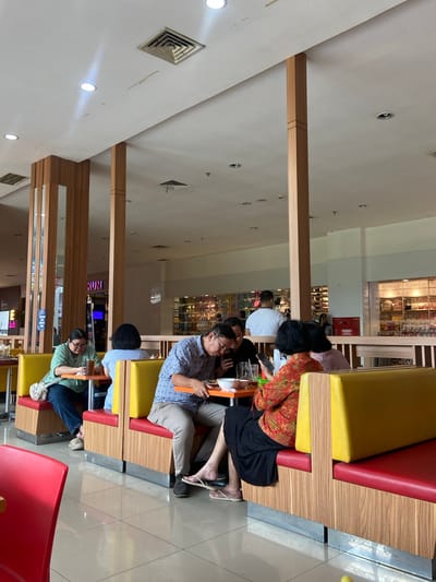The image shows a group of people sitting at tables in what appears to be a food court or restaurant. The setting is indoors with a tiled floor and a white ceiling with some visible vents and lights. Wooden pillars and partitions divide the space. In the foreground, several people are seated at booth-style tables with red and yellow upholstery. One person in a green shirt is looking down at a table with a drink. Next to them, a person in a blue and white patterned shirt is also looking down, possibly at their phone or food. Further to the right, a woman in a vibrant orange and red patterned top and a black skirt is looking at her phone. Behind them, more people are visible, and in the background, there are displays of merchandise, suggesting a connection to a shopping mall. One sign in the background reads "RUN," possibly part of a store name. The overall atmosphere appears to be casual and relaxed, with people engaged in dining and personal activities. The image quality suggests it was taken with a mobile phone.