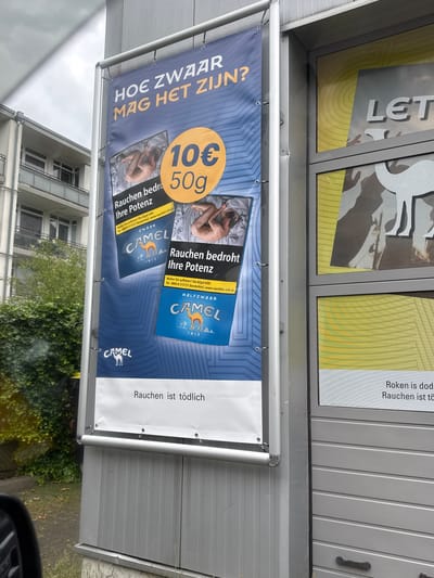 The image shows an outdoor advertisement billboard in Gronau, Germany. The billboard features a large blue banner with text in Dutch and images of Camel cigarette packs. The main text on the banner reads "HOE ZWAAR MAG HET ZIJN?" which translates to "HOW HEAVY CAN IT BE?". Below this, there are images of Camel cigarette packs with the text "Rauchen bedroht Ihre Potenz" meaning "Smoking threatens your potency". A circular graphic displays "10€" and "50g", likely referring to the price and quantity of the cigarettes. The bottom of the billboard has the text "Rauchen ist tödlich" which means "Smoking is deadly".
To the right of the billboard, a store window is visible with a yellow and grey design. Part of the word "LE" is visible in large white letters on a grey background, possibly part of a store name. A white silhouette of a camel is also visible on the window.
The environment around the billboard includes a building with balconies and green ivy growing on it, suggesting an urban or suburban setting. The bottom of the image shows a car's side mirror and part of a car's interior, indicating the photo was taken from a vehicle. The overall lighting suggests it is daytime, with overcast skies.