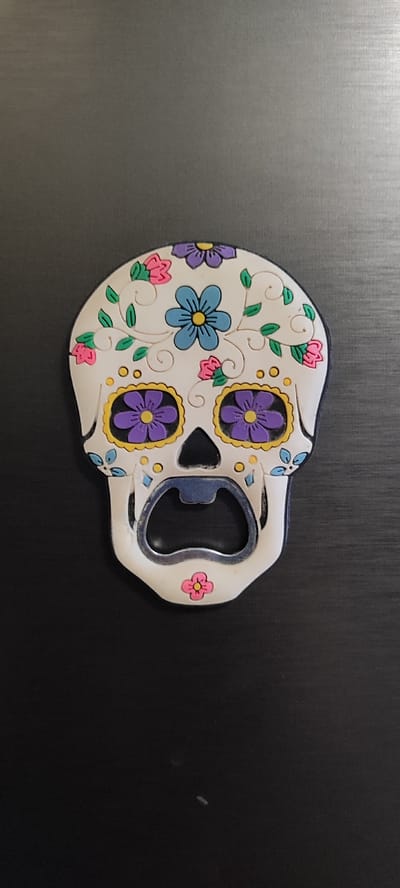 This media file features a close-up, overhead view of a sugar skull-shaped bottle opener resting on a dark, textured surface that appears to be wood grain. The bottle opener is predominantly white with colorful decorations in a style reminiscent of Dia de los Muertos imagery.

The skull design is adorned with various floral elements. There is a blue flower with a yellow center on the forehead, and two large eye sockets are decorated with purple flowers with yellow centers, each outlined in yellow. Smaller pink and blue flowers, along with green leaves and vines, are scattered across the top of the skull. Tiny yellow circles are also part of the decorative pattern.

The metallic part of the bottle opener, which is exposed in the jaw area of the skull, has a silver finish and shows some signs of wear. A small pink flower is depicted at the very bottom of the skull, below the bottle opener mechanism.

The background is a uniform dark color with a subtle horizontal texture, likely a wooden table or countertop. There are no visible people, animals, or specific location cues beyond the provided location context of Dinskaya, Russia, which cannot be determined from the image itself. The lighting suggests it is indoors, with the overhead shot creating soft shadows. No text is visible on the bottle opener or in the background. The overall mood is decorative and festive due to the vibrant colors and floral motifs on the skull.