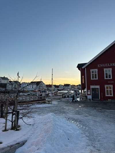 The image captures a detailed winter scene in Henningsvær, Norway, under a clear, transitioning sky with a pale blue upper half gradually shifting to a soft yellowish-orange glow at the horizon, indicative of low sun. The foreground features a snow and ice-covered ground, including a prominent mound of textured snow near the center-right. To the left, a small, bare-branched deciduous tree or shrub is visible, rooted near a dark stone curb.
On the right side of the midground stands a multi-story red wooden building with white window frames, trim, and eaves. The building's facade bears white lettering: "ENGELSK" across its upper section and "GLASS -" below. A dark-colored entrance door, marked with "No 1" above, is present on the ground level, flanked by several white informational signs on stands. A wooden ramp or walkway descends from the building towards the water. One individual, dressed in dark attire, is visible near the base of this ramp, appearing to be standing or moving towards the water.
The central and left portions of the midground are occupied by a harbor. Numerous boats, including several fishing vessels with masts and various smaller craft, are moored alongside wooden docks and jetties. Some boats show visible signs of snow or ice accumulation. The water in the harbor appears dark and calm. In the background, a cluster of white and light-colored buildings with dark roofs, typical of a coastal village, line the distant shoreline, extending into the soft light of the low sun. The overall scene conveys a cold, winter environment in a Nordic fishing village.