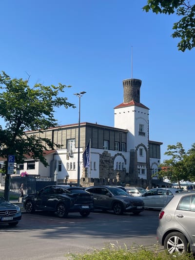In Wunstorf, Germany, a stone tower with a white facade, topped by a spire and a reddish-brown tiled roof, stands tall against a clear blue sky. The tower, bearing the inscription "Strandterrassen," is part of a building complex with white walls, stone accents, and wooden louvers on the upper story. Several cars are parked in front of the building, including a dark SUV and a silver sedan. A green tree with lush foliage shades the scene, and a street lamp is visible on the right side. The ground is paved, suggesting a public area or parking lot. A person is walking away from the viewer on the left side of the image. A blue parking sign is also present on the left.
