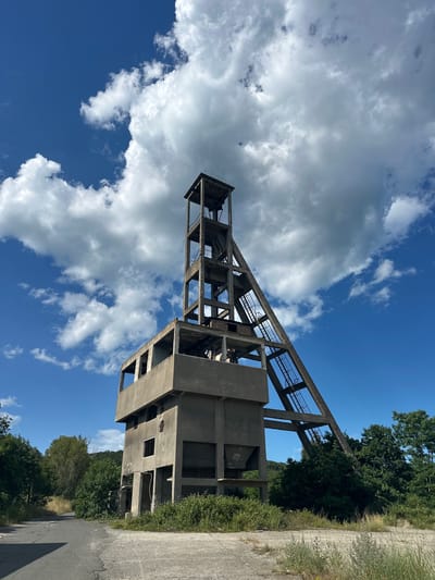 The image depicts an abandoned industrial structure, likely a mine headframe, situated in an outdoor environment under a bright blue sky with scattered white clouds. The structure is made of concrete and features a multi-story building at its base with several open window-like openings. Extending upwards and diagonally from the base is a tall, skeletal tower with exposed beams and a staircase or ladder system. Vegetation, including trees and grass, surrounds the structure and the paved, deteriorating road in the foreground. The scene suggests a disused industrial site, possibly in a rural or semi-rural setting. The location of Rosen, Bulgaria, is not explicitly visible within the image itself but is provided as context. There are no people or animals present in the image, and no actions or interactions are depicted. The environment is sunny and appears to be during the daytime.