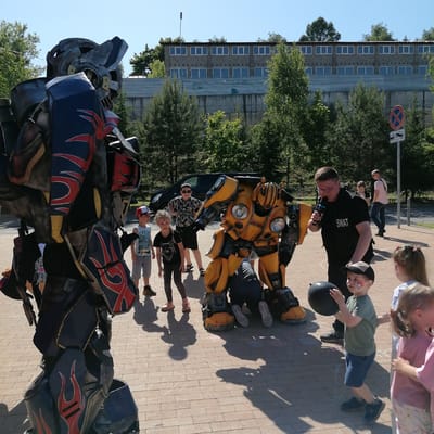This image captures an outdoor event in Трубачеевка, Russia, under clear skies and bright sunlight, casting distinct shadows on a light-colored brick paved surface.
In the foreground, a person in a large, dark-toned robot costume, resembling Optimus Prime with red and blue flame detailing on black and grey armor, stands on the left, partially cropped, facing right. To its right, centrally located, a person in a yellow and black robot costume, stylized as Bumblebee, is crouched or kneeling, positioned to interact at a lower height.
An adult male, wearing a black "SWAT" vest and clear glasses, stands to the right of the yellow robot, holding a black microphone to his mouth, appearing to address the gathered children. Several children are present: a boy in a white and red cap and blue t-shirt stands near the yellow robot, while a girl in a black t-shirt stands next to him. Behind them, a boy in sunglasses and a patterned shirt observes. In the immediate foreground on the right, a young boy with red face paint (a flame motif) and a dark baseball cap holds a black balloon, looking towards the entertainer. Two other young girls are partially visible to his right.
In the mid-ground, a black van is parked amidst trees. In the background, a large, two-story building constructed of grey concrete blocks with numerous rectangular windows is visible. Below this building, a corrugated metal roof section is present, followed by a concrete wall with faint graffiti. Various green trees are dispersed throughout the background and mid-ground. A tall, dark street light pole stands to the left of the building's upper section. A road sign on the right indicates "No Stopping/Standing" (red circle with blue X) and a supplementary sign showing a tow truck. The scene suggests a public performance or entertainment event for children.