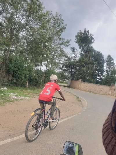 The image depicts an outdoor scene in Machakos Central ward, Kenya, on an overcast day. The central subject is a person riding or standing with a bicycle on a paved road. The individual is viewed from behind, wearing a red short-sleeved shirt with the white number "5" prominently displayed on the back, along with faint letters "KAN" or "KAI" above it. They are also wearing dark trousers, light-colored shoes, and a light-colored cap or head covering.
The bicycle appears to be a mountain bike with a dark frame and a yellow reflector on its rear fender. The road is asphalt, marked with a white line along its edge, and features an unpaved, earthy shoulder. It curves gently to the right, leading into the distance. The surrounding environment is lush with vegetation, including numerous tall trees and dense bushes lining both sides of the road. A stone or brick wall is partially visible through the trees on the right side of the road. The sky is uniformly grey and cloudy, indicating an overcast daytime. In the upper right corner, a portion of a vehicle's side mirror is visible, suggesting the photograph was taken from another vehicle.
